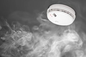 A smoke detector mounted on a ceiling with wisps of smoke curling around it, indicating it's detecting smoke or fire.