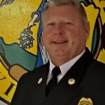 A smiling man in a formal black uniform with badge, posing in front of a colorful mural featuring a lion.