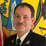 A man, dressed in a dark formal uniform with gold insignia, stands in front of a brightly colored background featuring a partial view of an American flag and another emblem with a blue and white figure. He has short dark hair, a mustache, and is smiling softly.