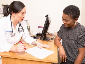 A female doctor in a white lab coat consults with a female patient, both smiling and looking at a document on the desk in a medical office.