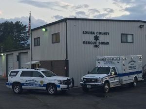 An ambulance and a police suv parked in front of the louisa county rescue squad building during twilight. the american flag flies near the building.