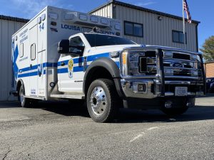 A blue and white louisa county ambulance parked in front of a building, prominently displaying the logo and emergency number, with an american flag in the background.