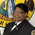 A smiling woman in a police uniform standing in front of a colorful emblem featuring an eagle and a shield.