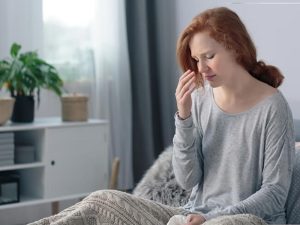 A woman with red hair, looking unwell, sits on a bed covered with a blanket, holding a tissue and pinching the bridge of her nose, with indoor plants and shelves in the background.