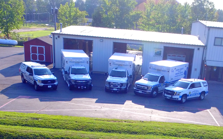 A lineup of emergency medical service vehicles is parked outside a building. The fleet includes several large emergency trucks and smaller SUVs, all marked with medical and emergency logos. Trees and additional buildings are visible in the background.